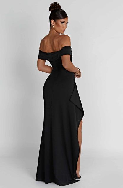 Back three-quarter view of the black off-shoulder pleated maxi dress, highlighting the high slit and draped accent.