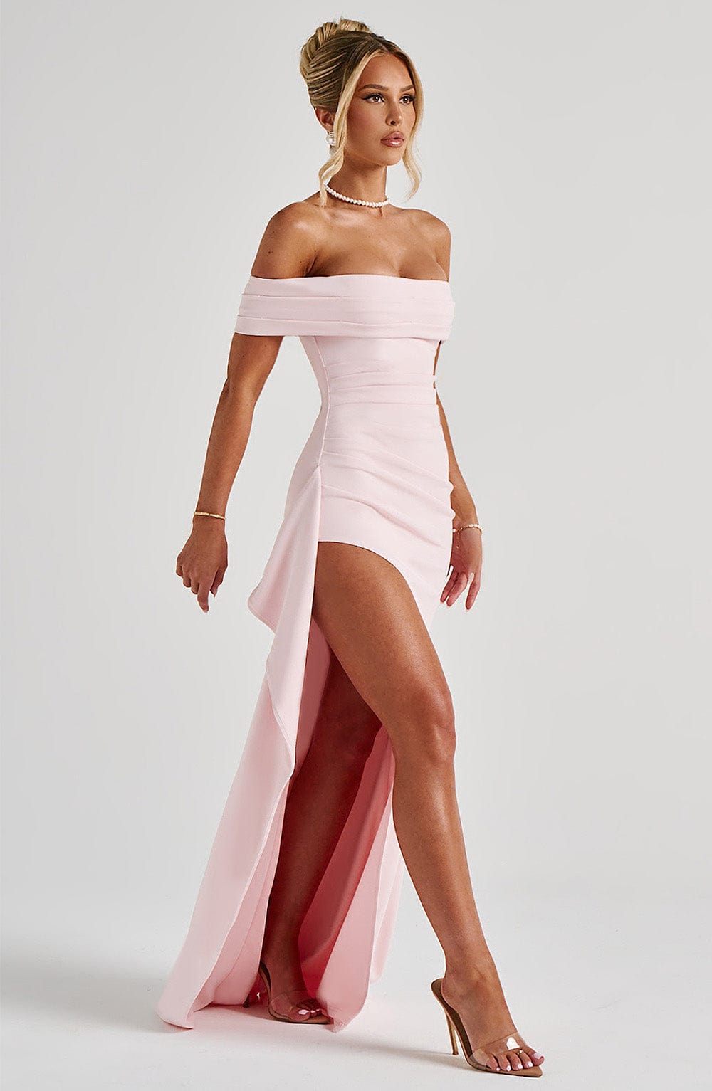 Side view of a light pink off-shoulder pleated maxi dress featuring a dramatic high slit and elegant draping.