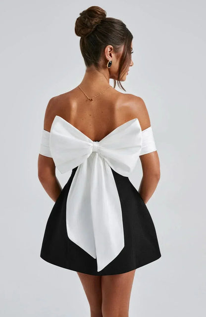 Back view of women's black mini dress featuring a large, white detachable bow and off-shoulder neckline.