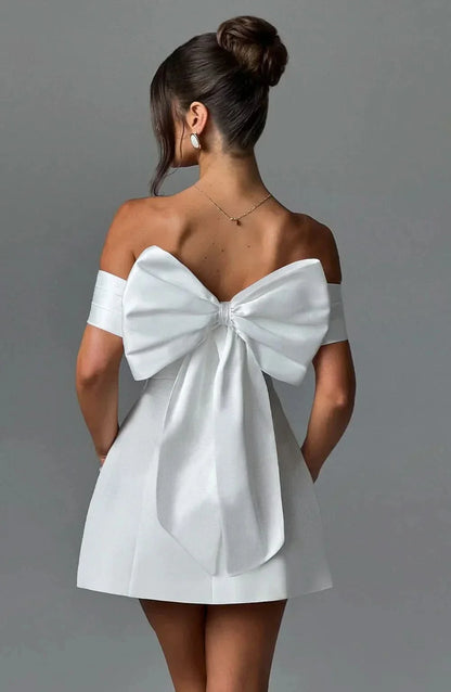 Back view of women's white mini dress with a large, elegant detachable bow and off-shoulder design.