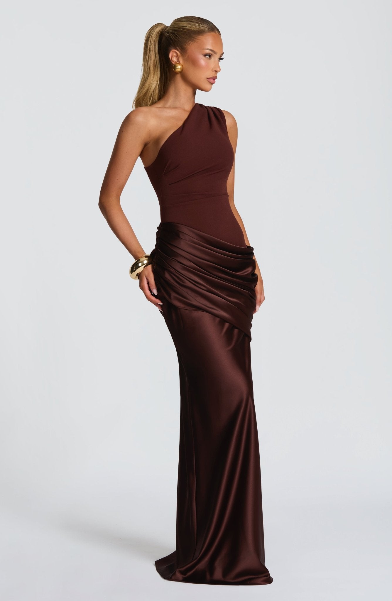 Side profile of a women's brown one-shoulder formal maxi dress with elegant draping, styled with a gold cuff bracelet.