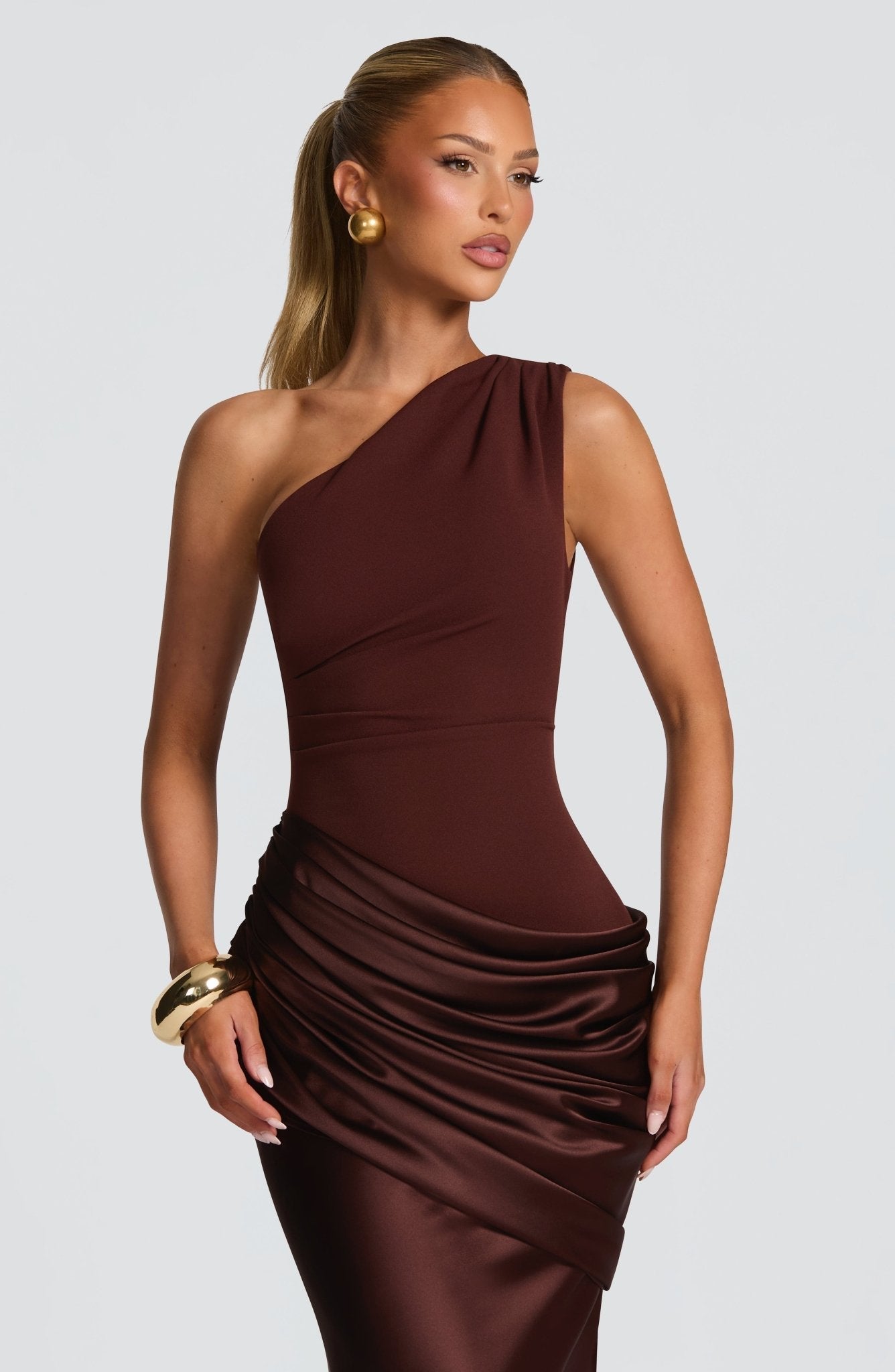Close-up of the bodice and draped waist of a women's chocolate brown one-shoulder formal maxi dress, with gold earrings and bracelet.
