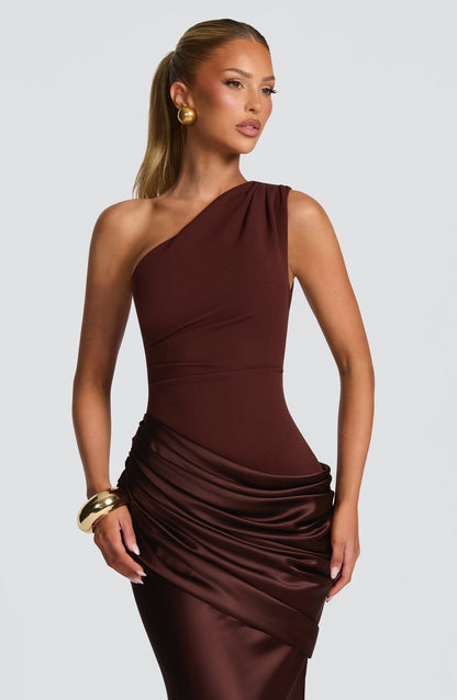 Close-up of the bodice and draped waist of a women's chocolate brown one-shoulder formal maxi dress, with gold earrings and bracelet.