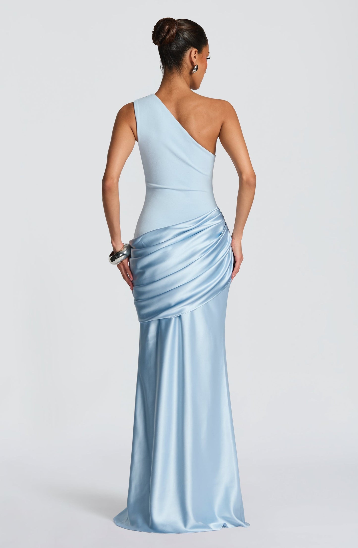 Back view of a women's light blue one-shoulder draped formal maxi dress, showcasing the graceful flow of the skirt.