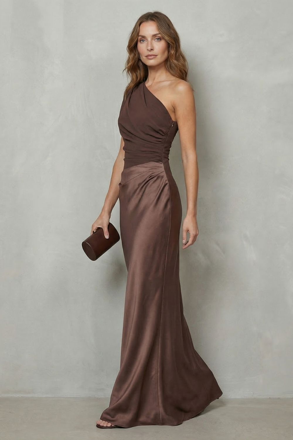 Side view of the women's brown one-shoulder draped satin maxi dress, highlighting the gentle drape of the skirt and a dark clutch.