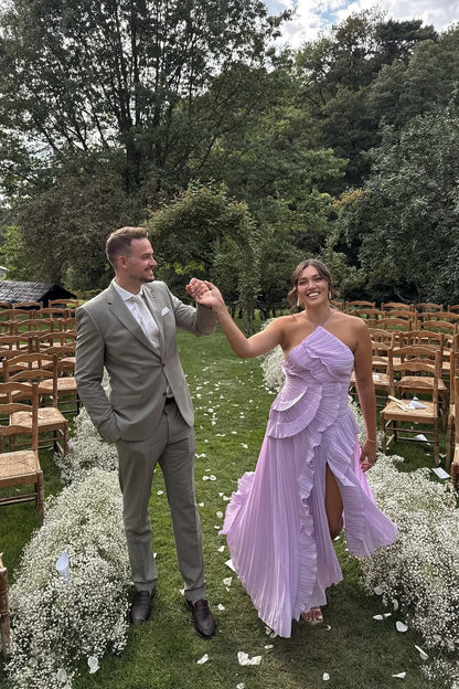 Woman in a pink one-shoulder ruffle pleated midi dress walking with a man in a grey suit at an outdoor event.