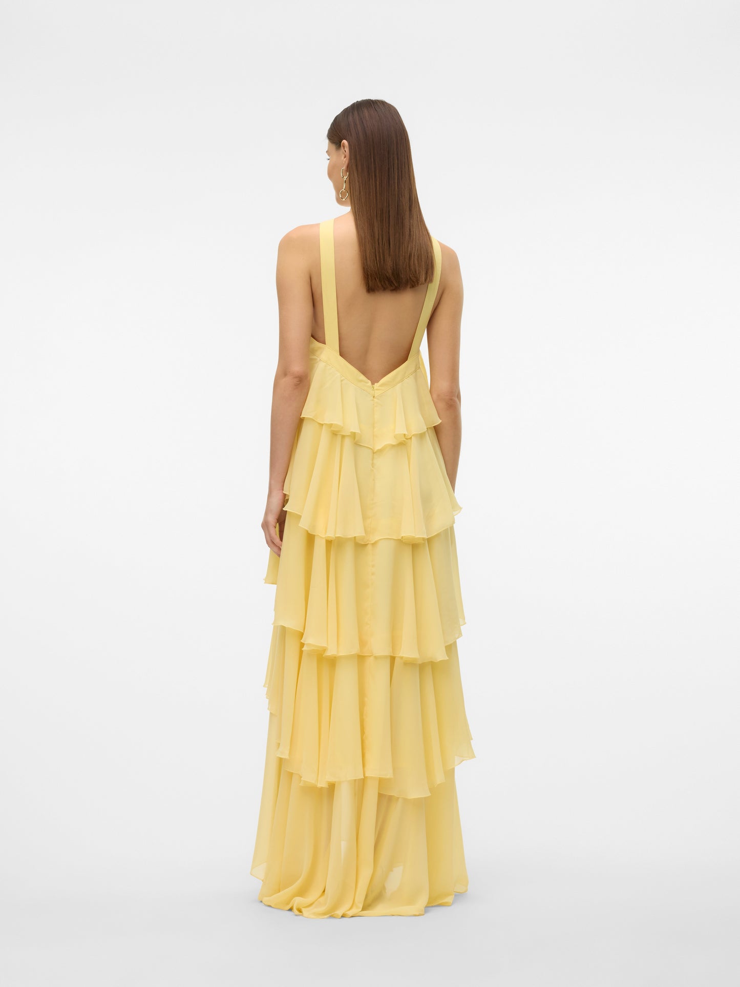 Full-length view of a woman wearing a flowing yellow halter ruffle backless maxi dress with tiered ruffles and a rose detail at the neckline, paired with cream heels. Ideal for vacation dresses.