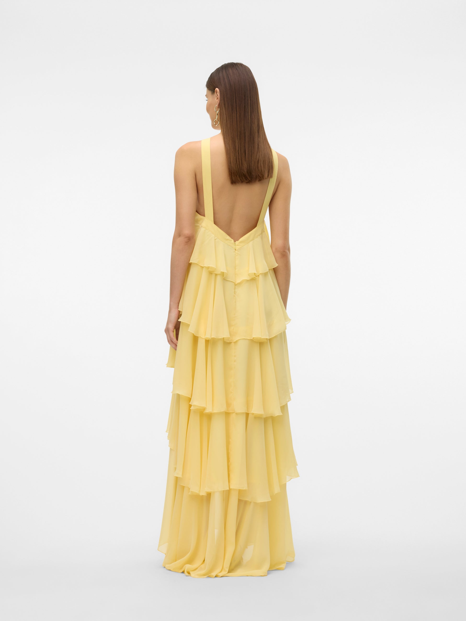 Full-length view of a woman wearing a flowing yellow halter ruffle backless maxi dress with tiered ruffles and a rose detail at the neckline, paired with cream heels. Ideal for vacation dresses.