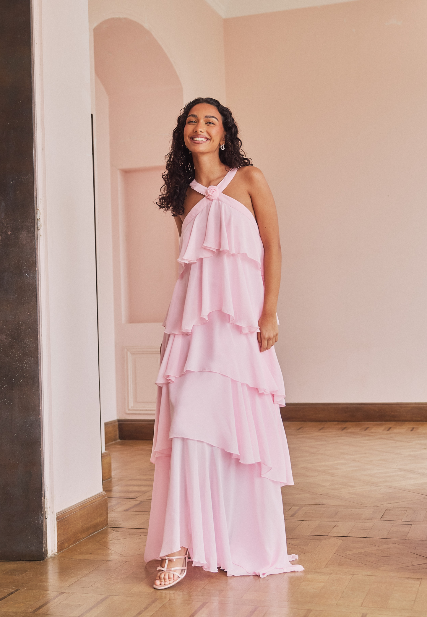 Smiling woman in a light pink tiered ruffle maxi dress with a halter neckline, perfect for wedding guests or vacation outfits.