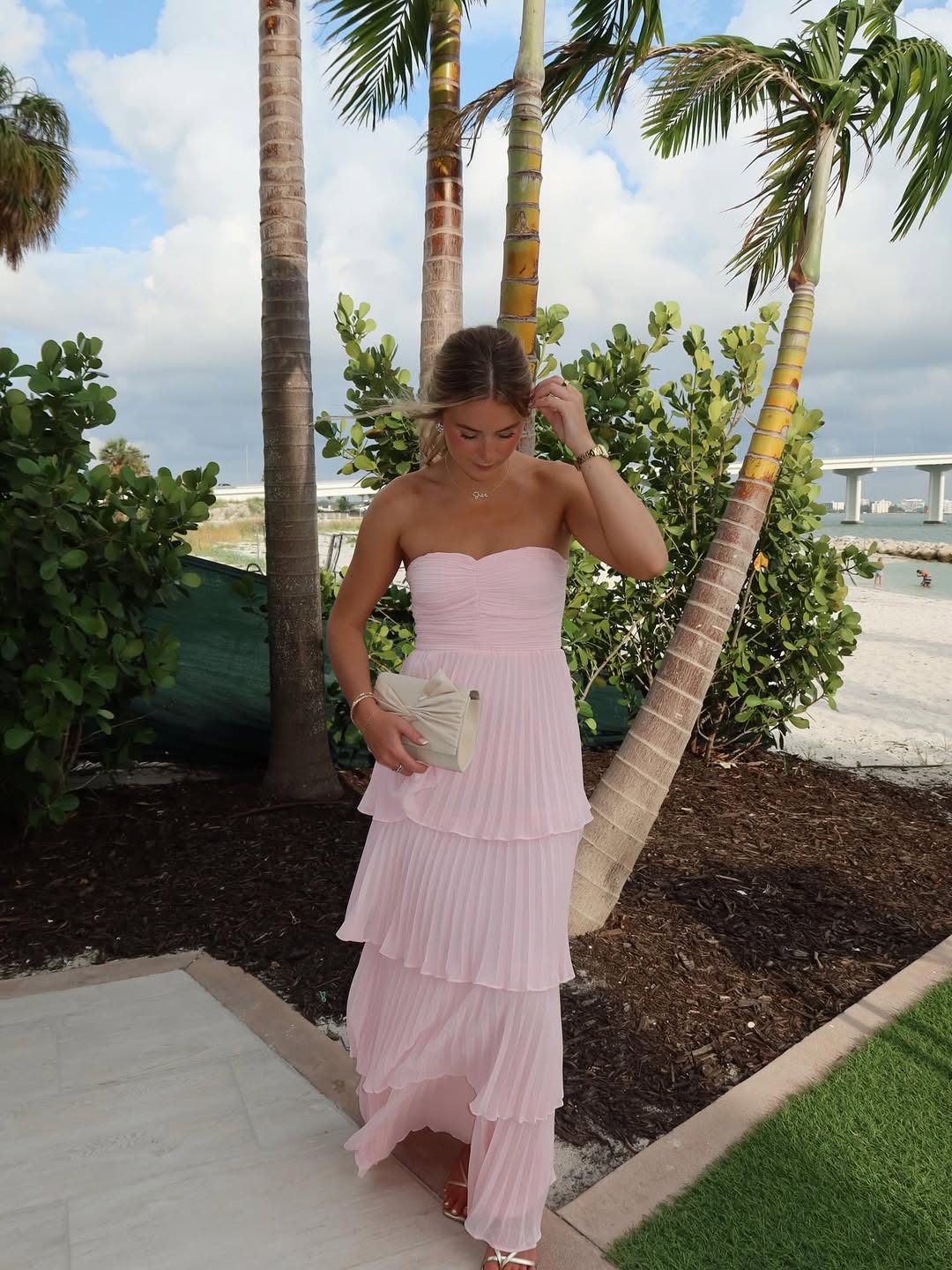 Light pink strapless tiered maxi dress with pleated skirt and white clutch, worn outdoors near palm trees, a great beach dress