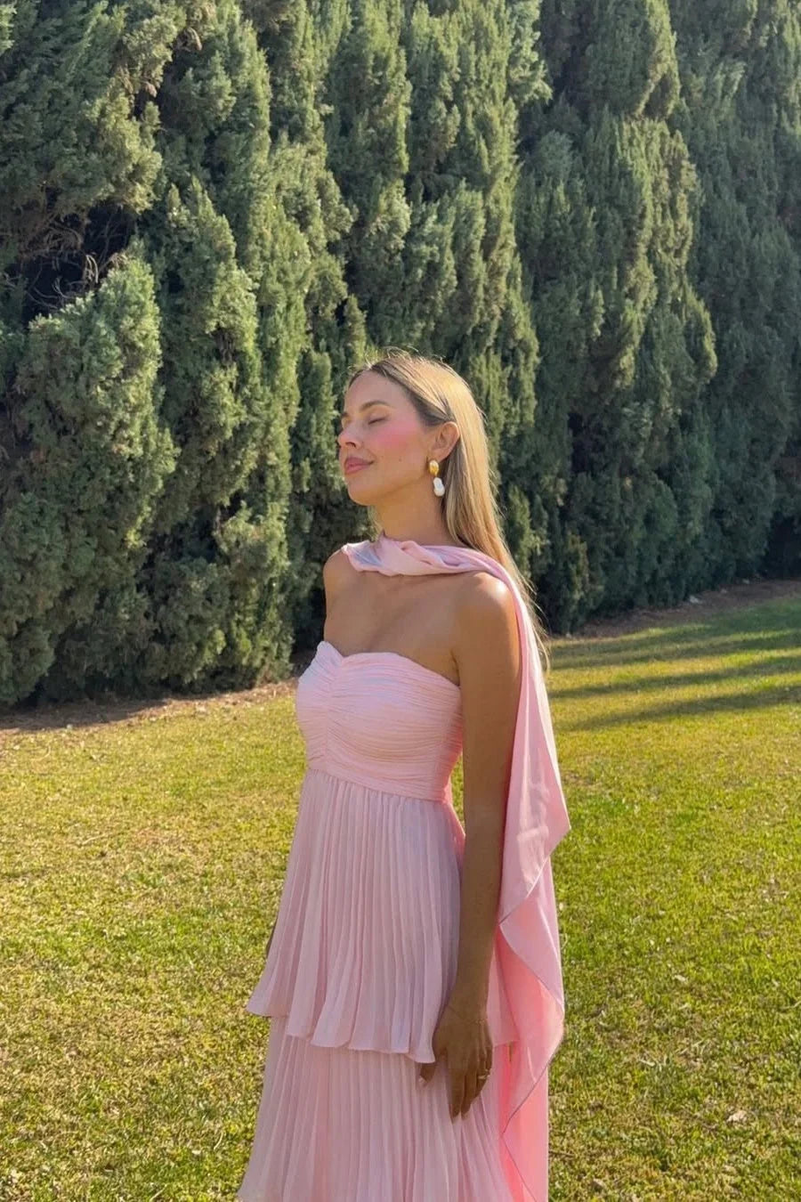 Elegant light pink strapless tiered maxi dress with pleated skirt and draped scarf, ideal for a vacation dress