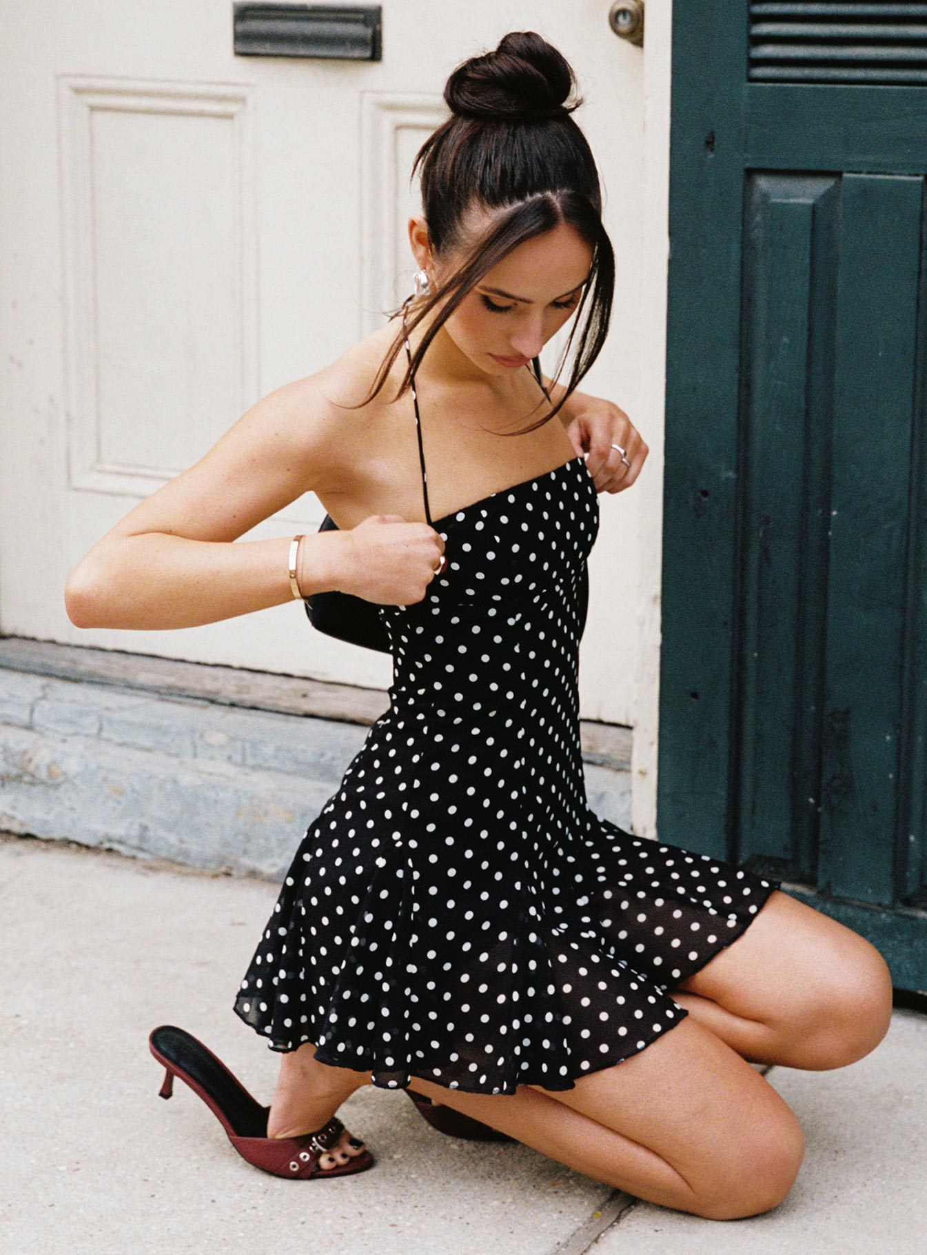 Women's black polka dot halter low back mini dress, kneeling outdoors, adjusting the halter straps.