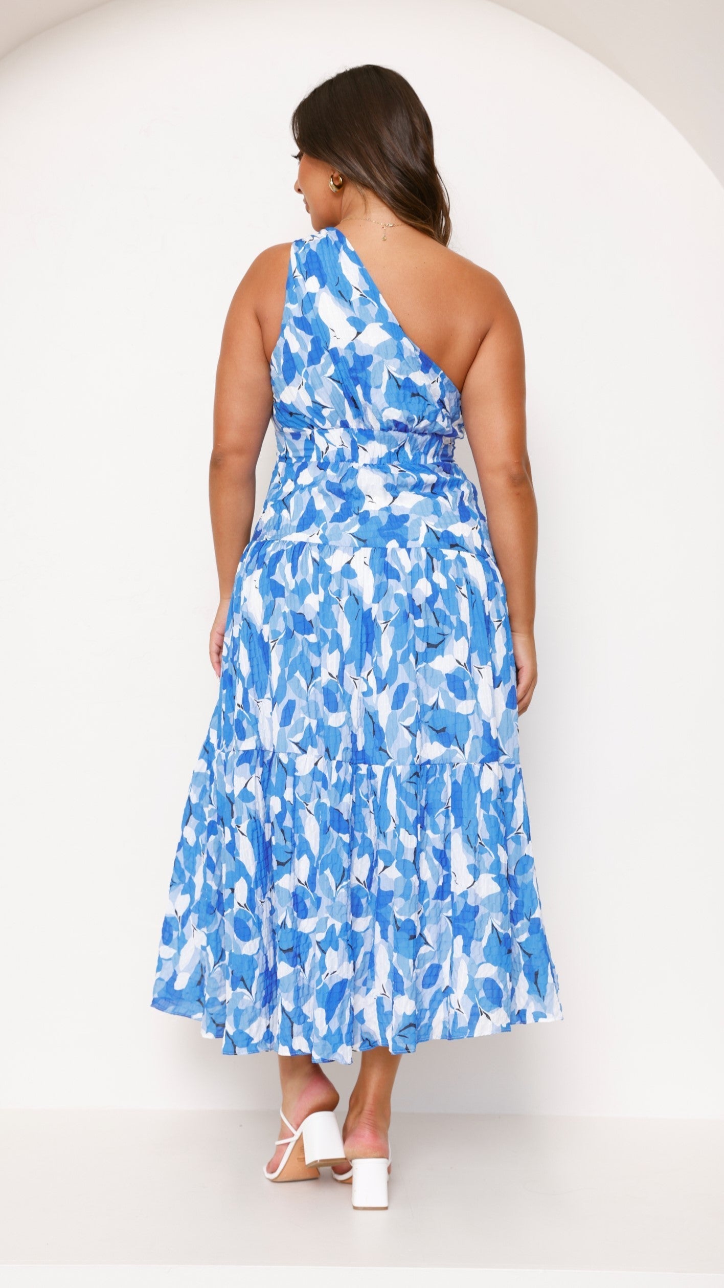 Back view of a plus size women's blue and white printed one-shoulder midi dress with a flowing tiered skirt.