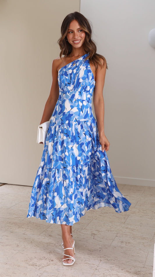 Smiling woman in a blue and white printed one-shoulder tiered midi dress, holding a white clutch and wearing white heels.