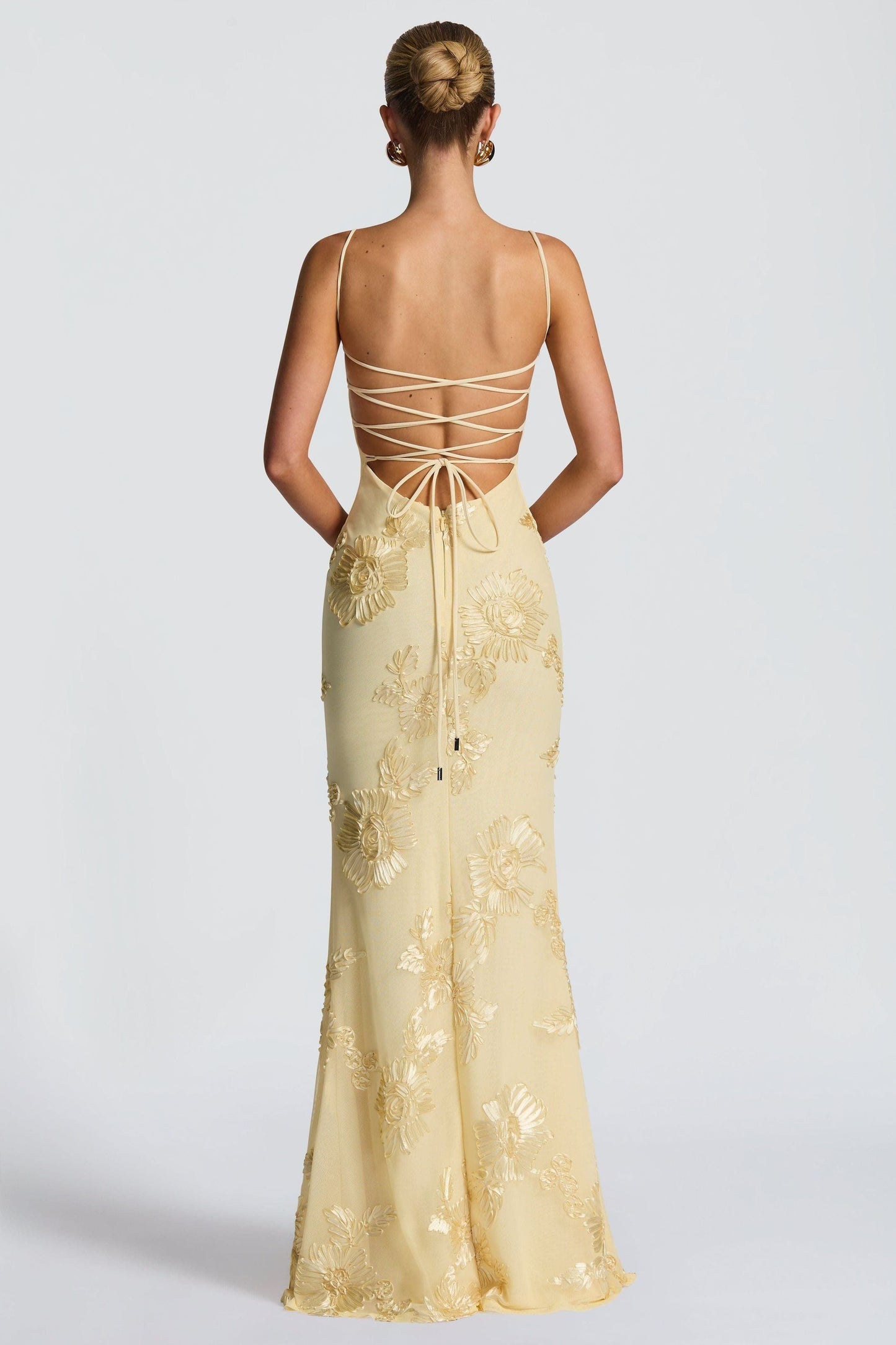 Outdoor shot of a woman in a light yellow floral sweetheart maxi dress leaning against a wall, showcasing the full length and design.
