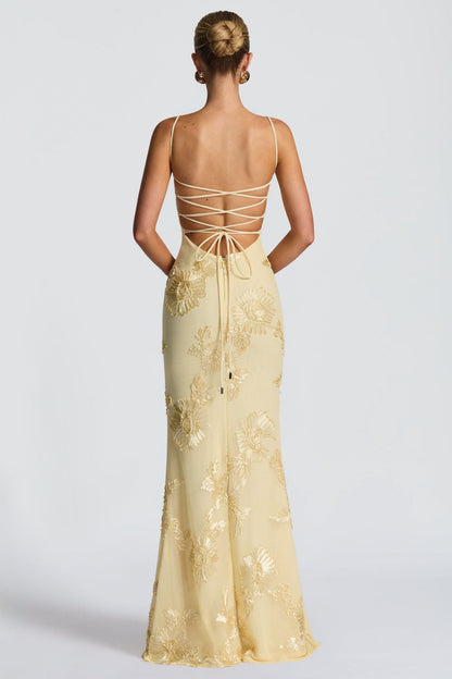 Outdoor shot of a woman in a light yellow floral sweetheart maxi dress leaning against a wall, showcasing the full length and design.