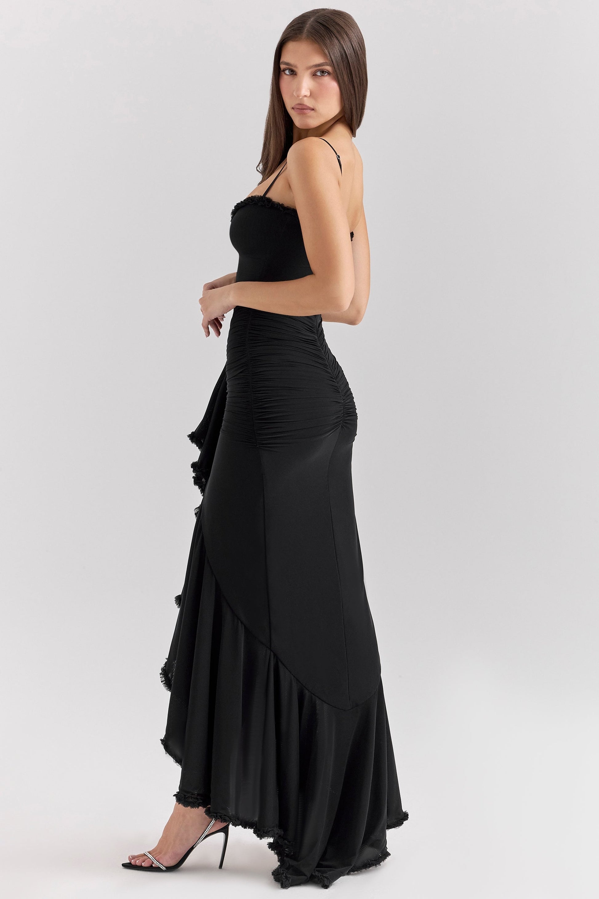 Side profile of a woman wearing a black sleeveless ruched ruffle high-low maxi dress, featuring a dramatic ruffled hem.