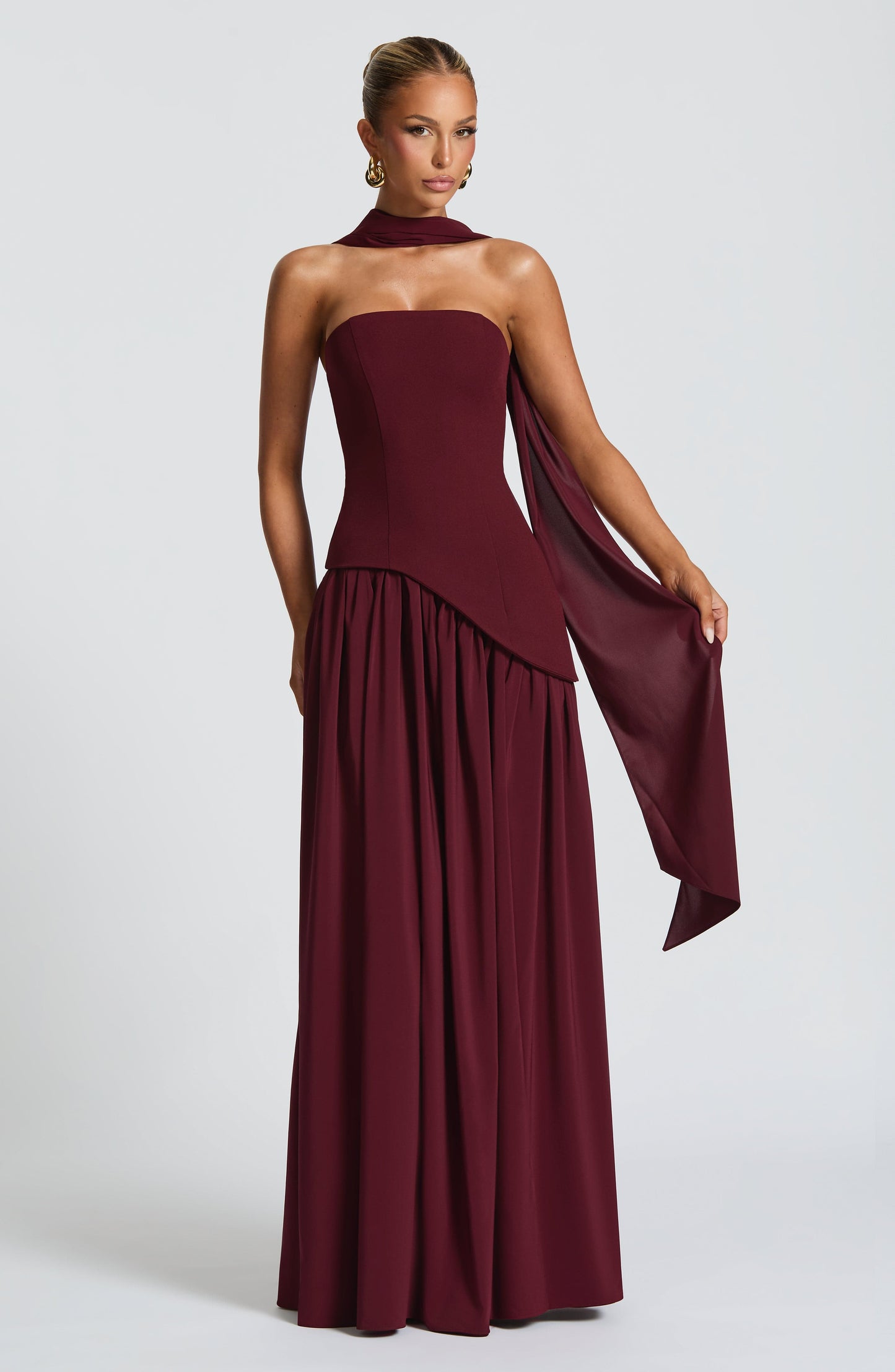 Front view of a women's strapless asymmetric corset maxi dress in deep burgundy, with a matching neck scarf and flowing fabric detail.