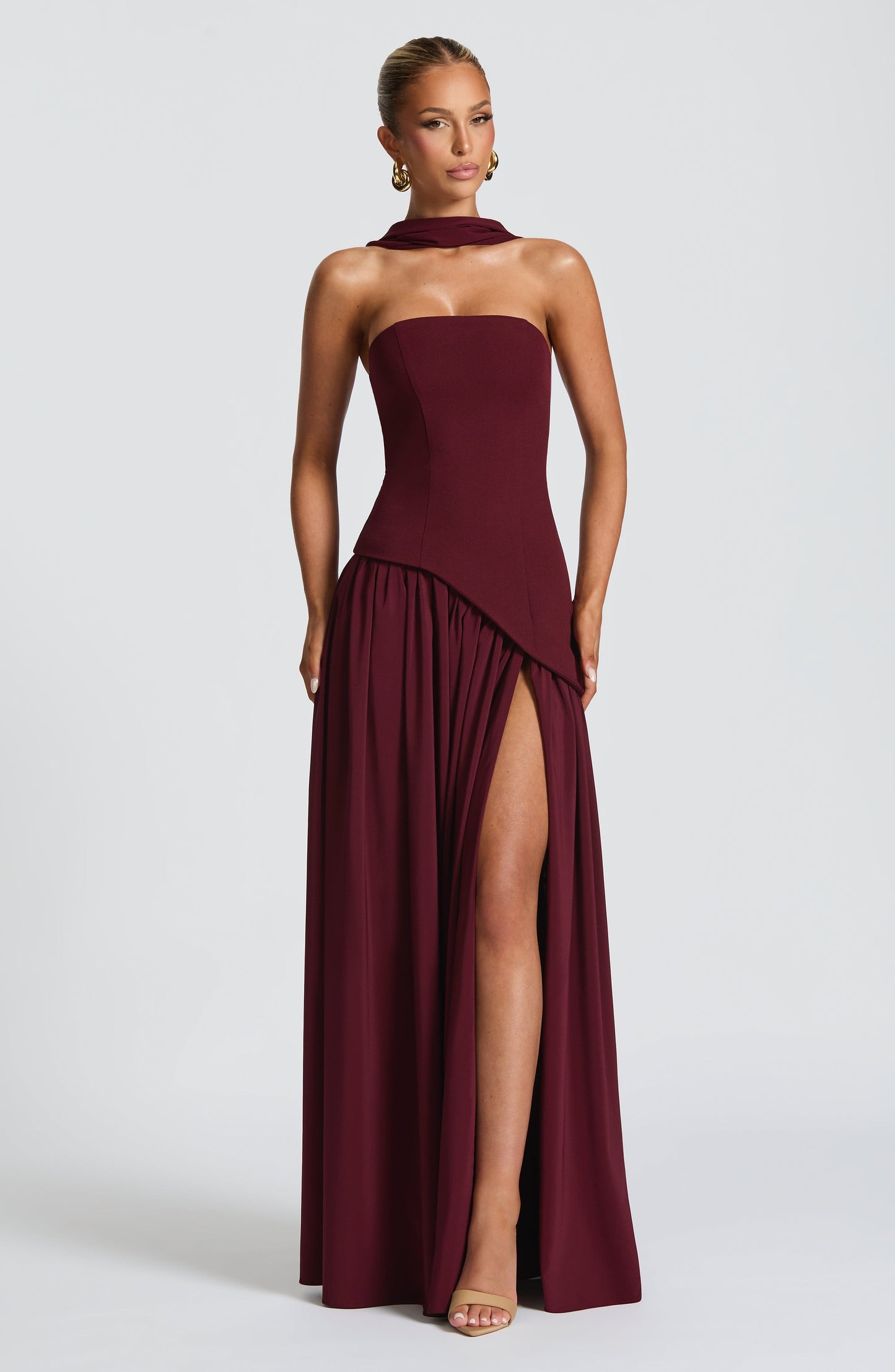 Full-length front view of a women's strapless asymmetric corset maxi dress in rich burgundy, showcasing a high leg slit.