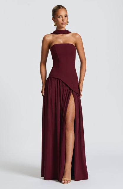Full-length front view of a women's strapless asymmetric corset maxi dress in rich burgundy, showcasing a high leg slit.
