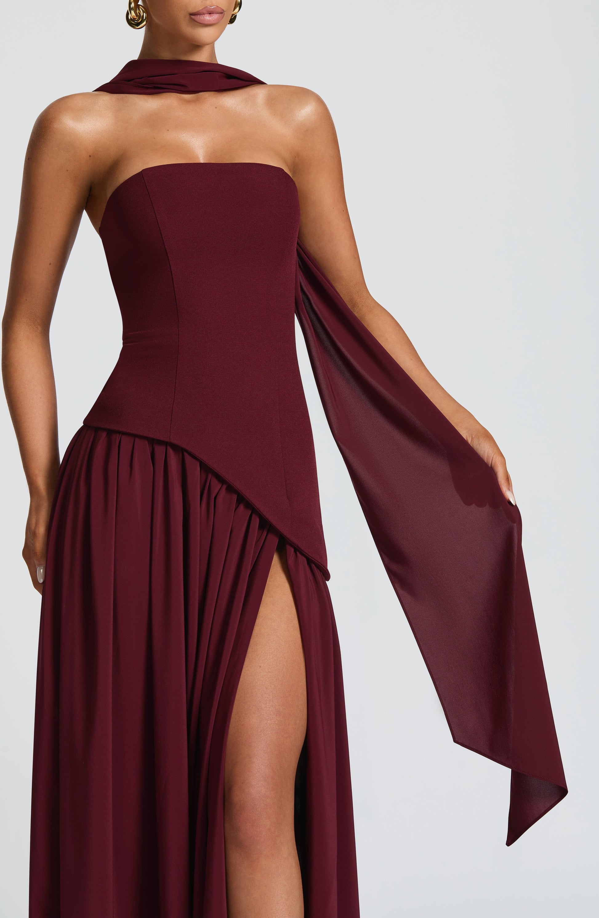 Close-up front detail of a women's strapless asymmetric corset maxi dress in burgundy, highlighting the bodice, flowing scarf, and leg slit.