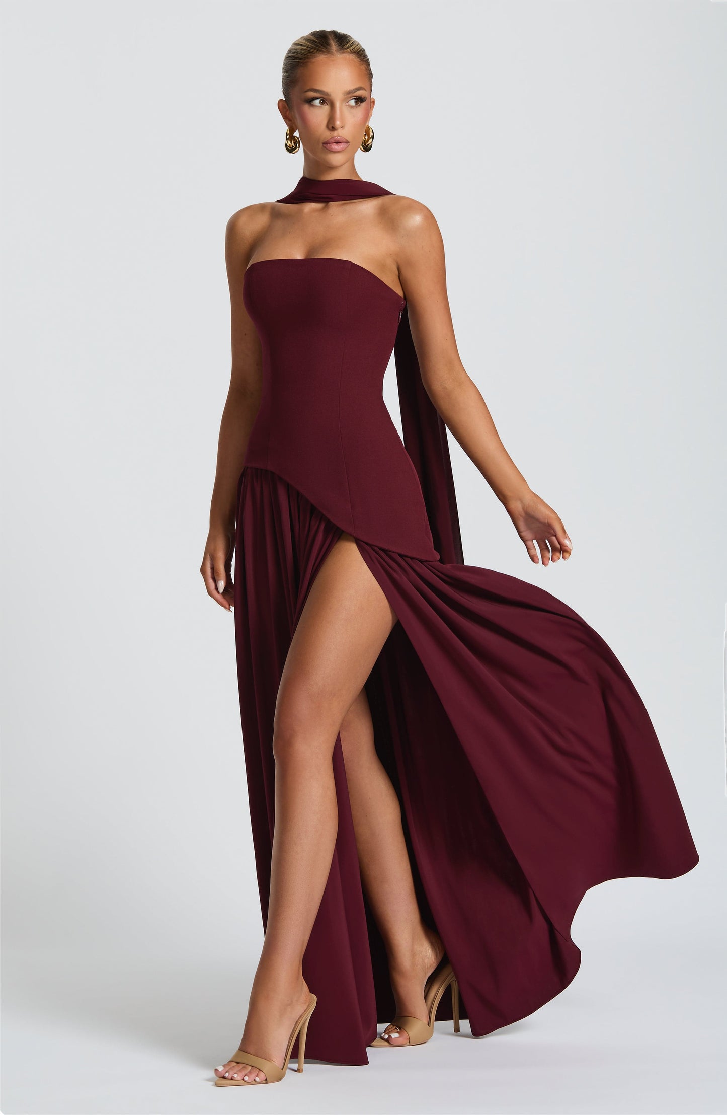 Dynamic front view of a women's strapless asymmetric corset maxi dress in burgundy, with the skirt flowing and a high leg slit visible.