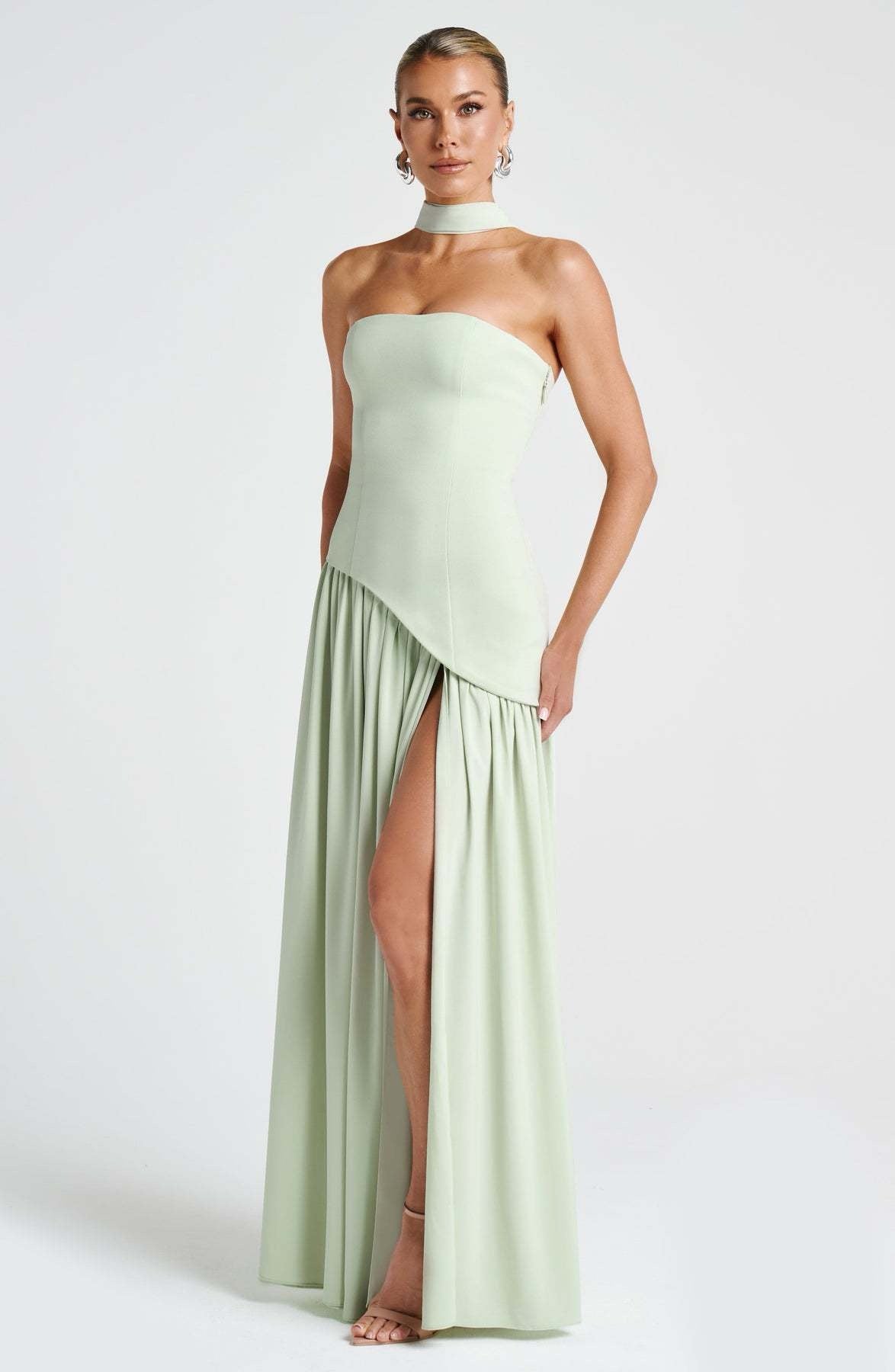 Full-length front view of a women's strapless asymmetric corset maxi dress in light sage green, highlighting the choker and high leg slit.