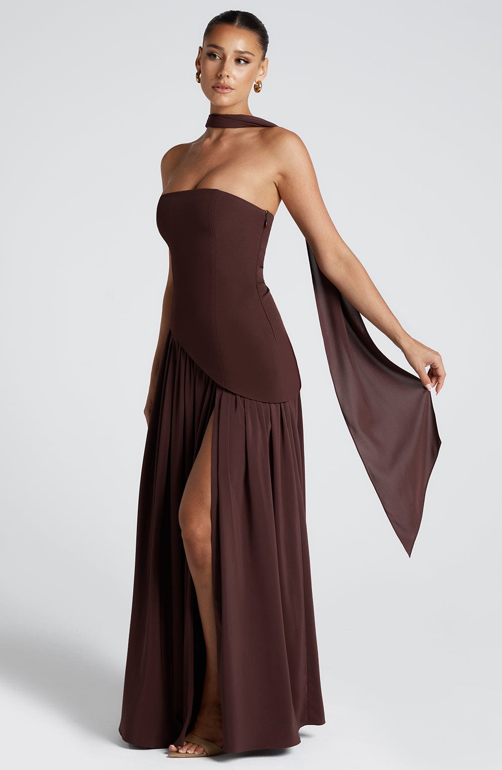 Side view of a brown strapless asymmetric corset maxi dress featuring a thigh-high slit and elegant scarf.