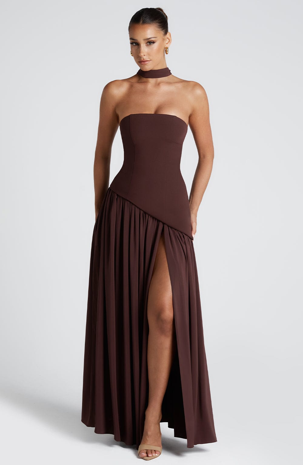 Full front view of a brown strapless maxi dress with a fitted corset bodice, asymmetric hem, and high leg slit.