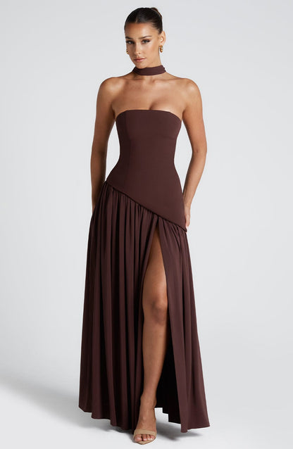 Full front view of a brown strapless maxi dress with a fitted corset bodice, asymmetric hem, and high leg slit.