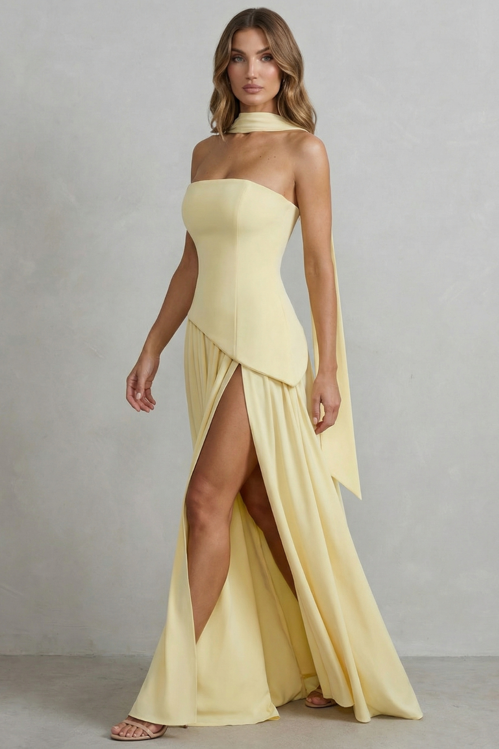 Model in a pale yellow strapless asymmetric maxi dress with a high leg slit and a long flowing scarf detail, perfect for vacation outfits.