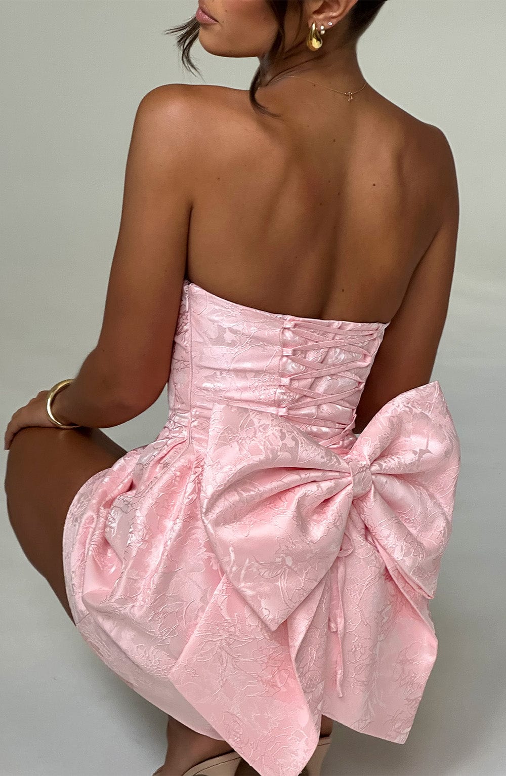 Close-up back view of the light pink floral mini dress on a model crouching, highlighting the large bow and lace-up back.