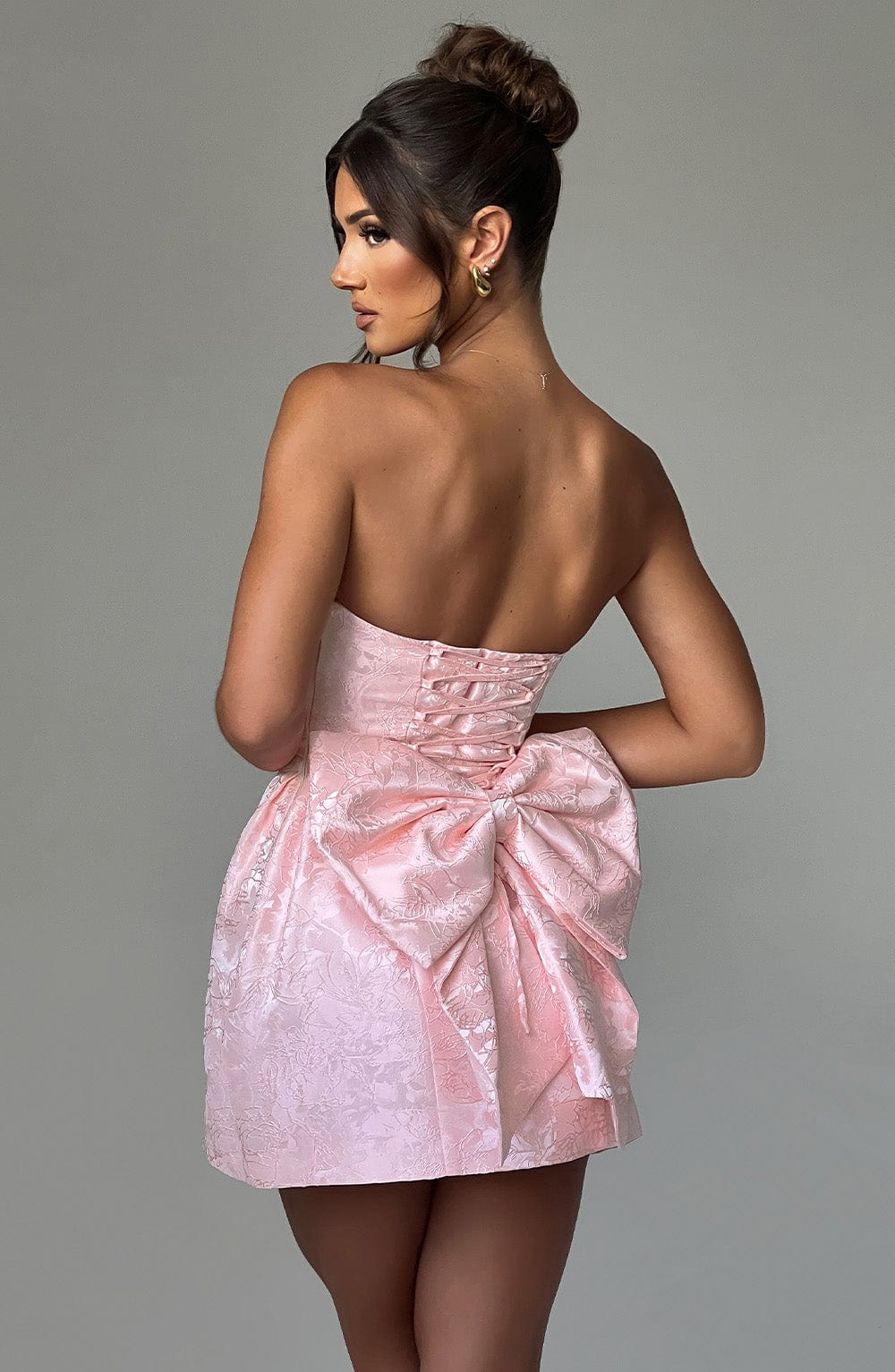 Three-quarter back view of the women's strapless pink floral mini dress with a large bow, showing the model looking over her shoulder.