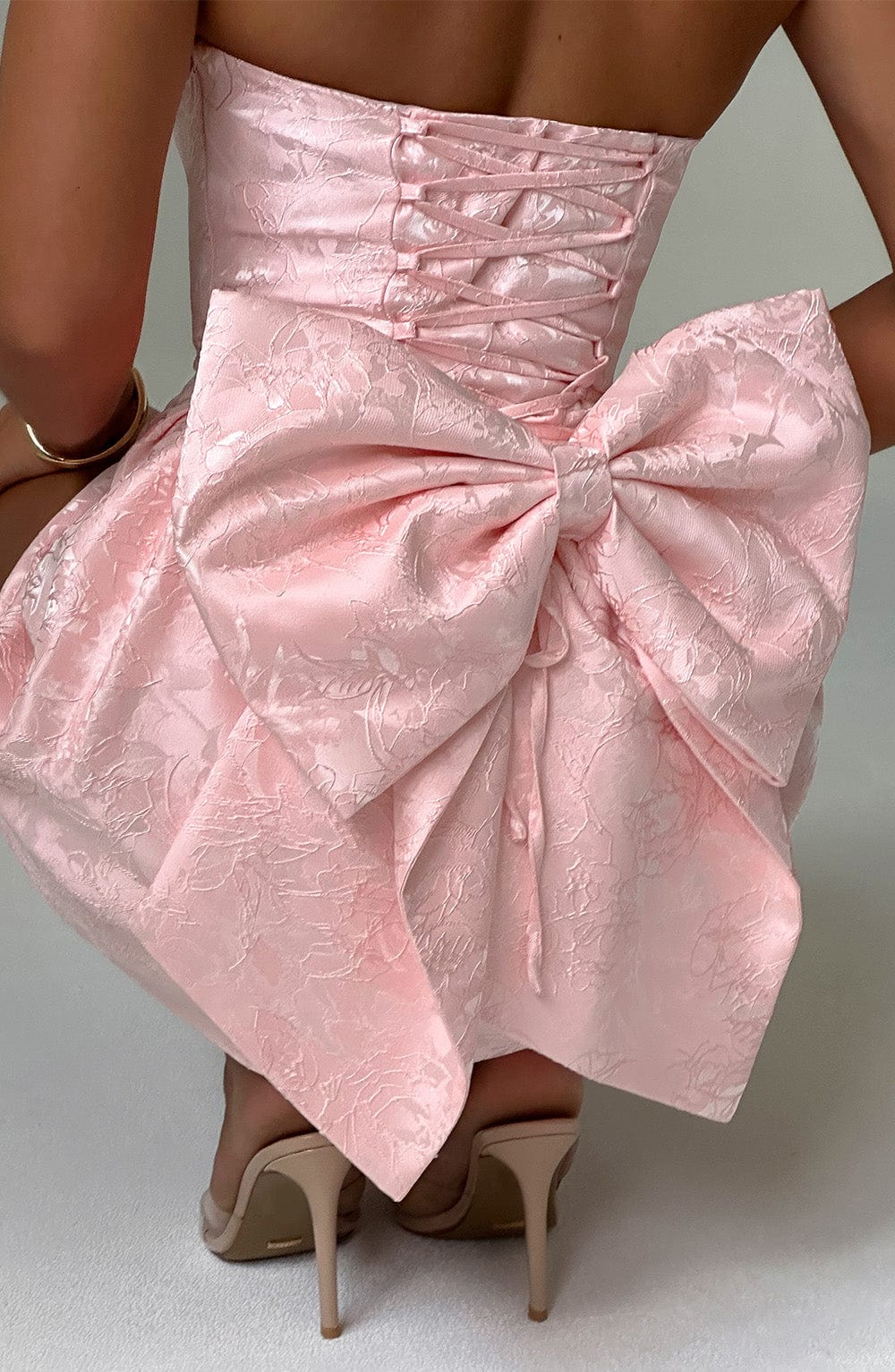Detailed back view of the light pink floral mini dress, focusing on the large bow, corset lacing, and nude high heels.