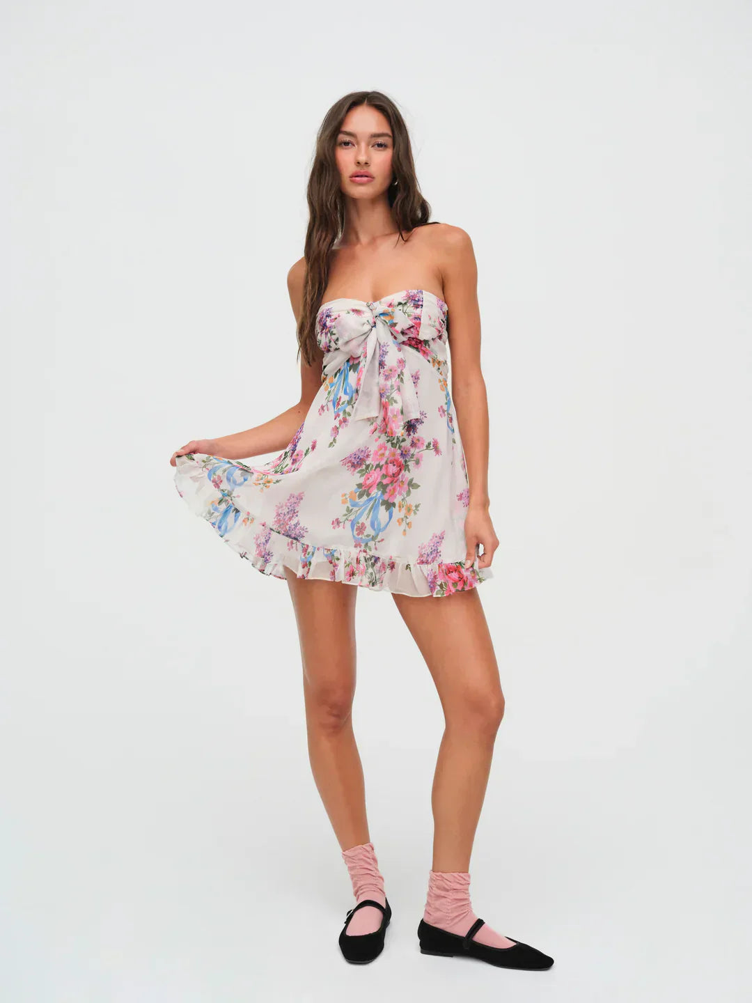 Full body shot of a women's strapless floral mini dress, white with pink, purple, and blue flowers, a ruffled hem, and a knotted bust, perfect for vacation outfits.