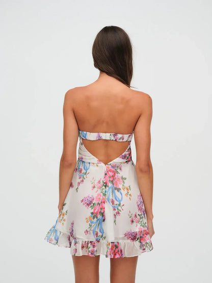 Back view of a women's strapless floral mini dress with an open-back design, showcasing the white fabric with pink and purple flowers and a ruffled hem.
