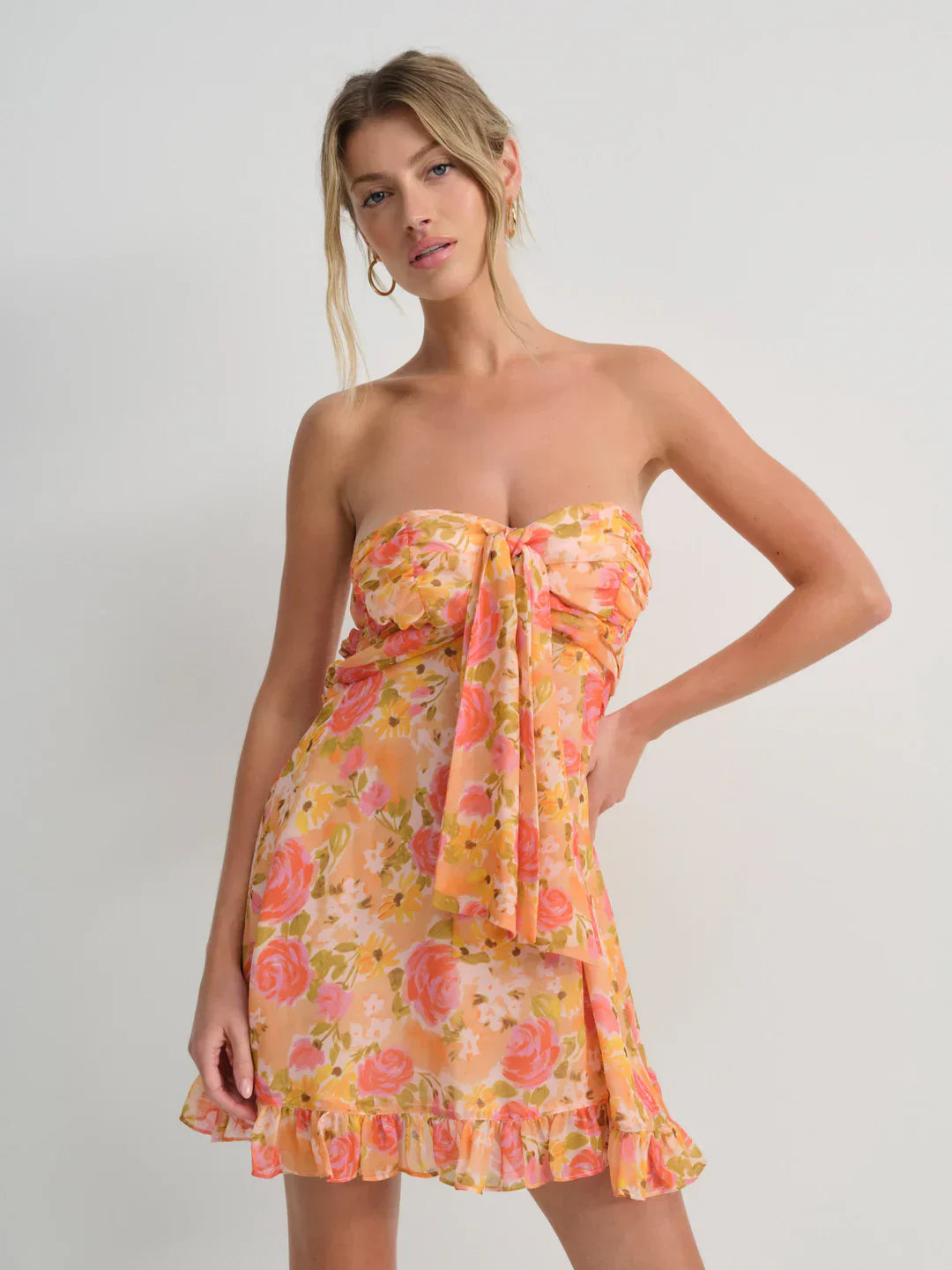 Front view of a women's strapless floral mini dress in a warm orange and pink floral print, featuring a knotted bust and ruffled hem, suitable for vacation outfits.