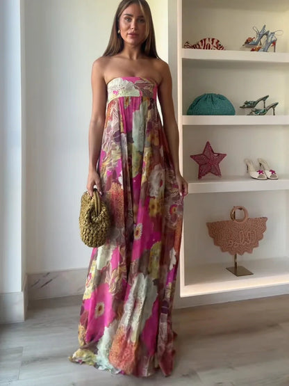Full-length women's strapless floral printed maxi dress in vibrant pink and orange tones, with a flowing silhouette, paired with a woven handbag.