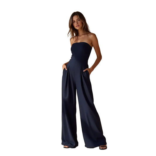 Full-body shot of a woman modeling a dark navy strapless high-waisted wide-leg jumpsuit with pleated details and pockets, against a beige background with shadows.