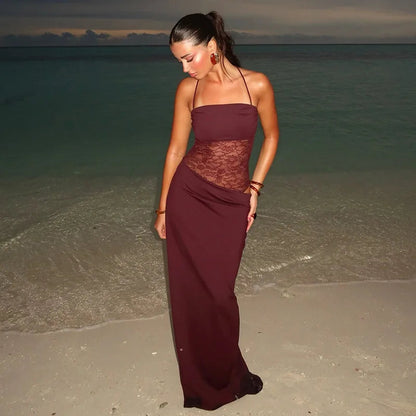 Front view of a woman wearing a burgundy strapless lace cut out maxi dress with thin halter straps, standing on a beach at dusk.