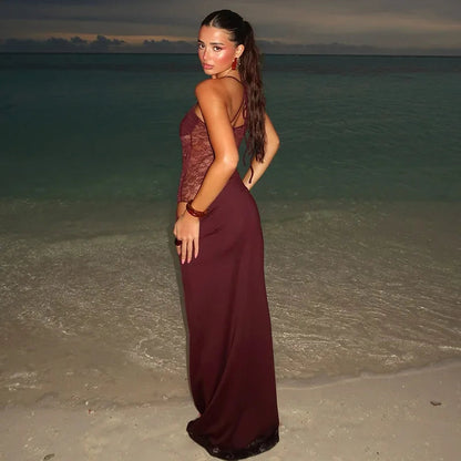 Back view of a woman in a burgundy strapless lace cut out maxi dress with thin halter straps, looking over her shoulder on a beach.