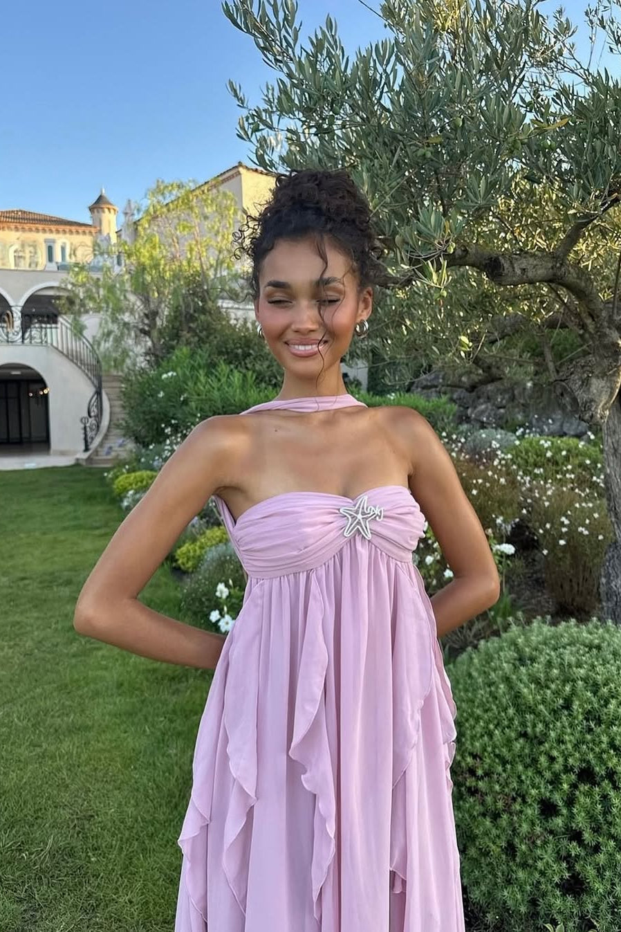 Women's light pink strapless ruched ruffle maxi dress with a starfish brooch, shown in a garden setting.