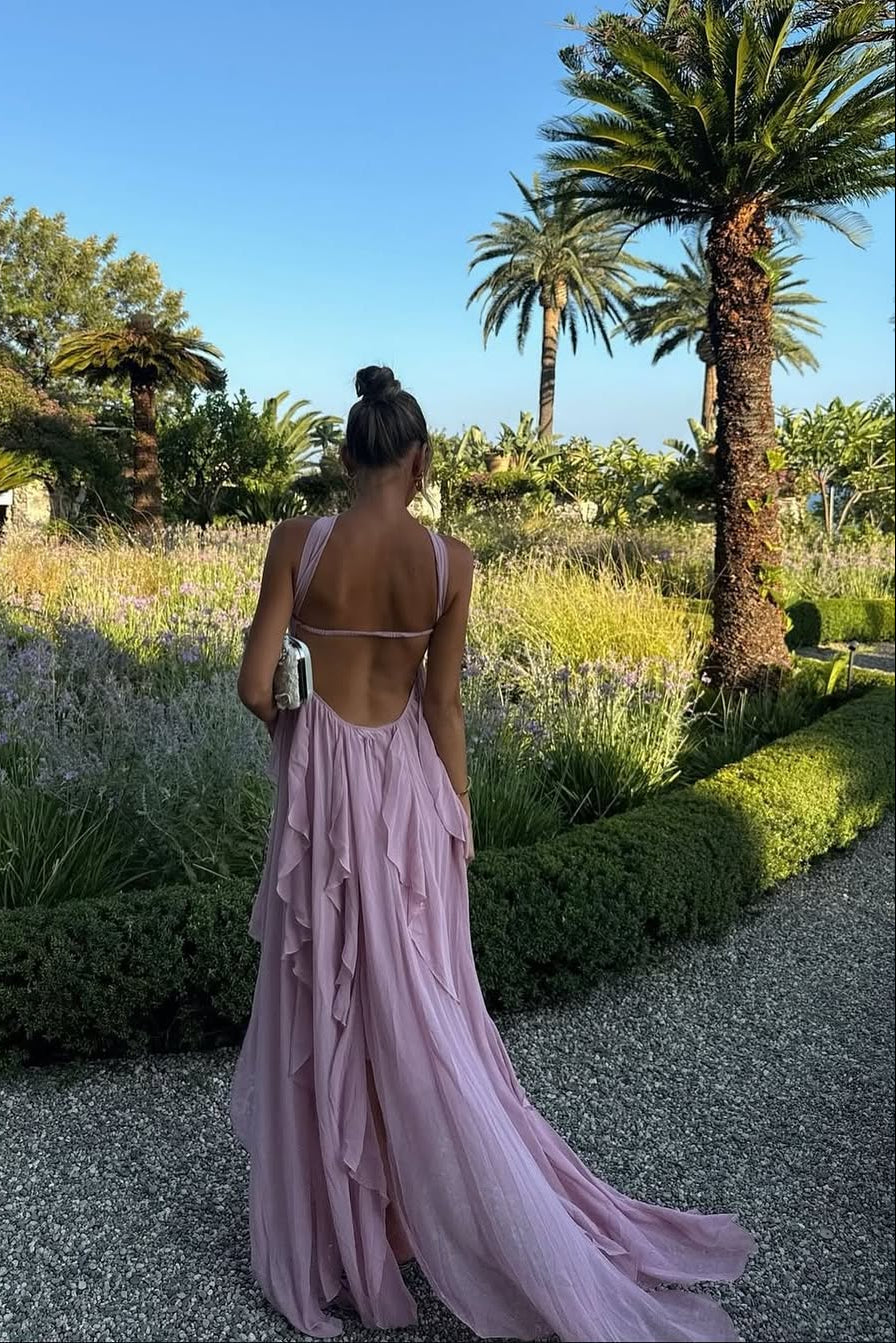 Back view of a women's light pink strapless ruched ruffle maxi dress with an open back and flowing skirt, in a garden.