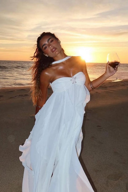 White strapless ruched ruffle maxi dress with a starfish brooch and choker detail, holding a wine glass on a beach at sunset.