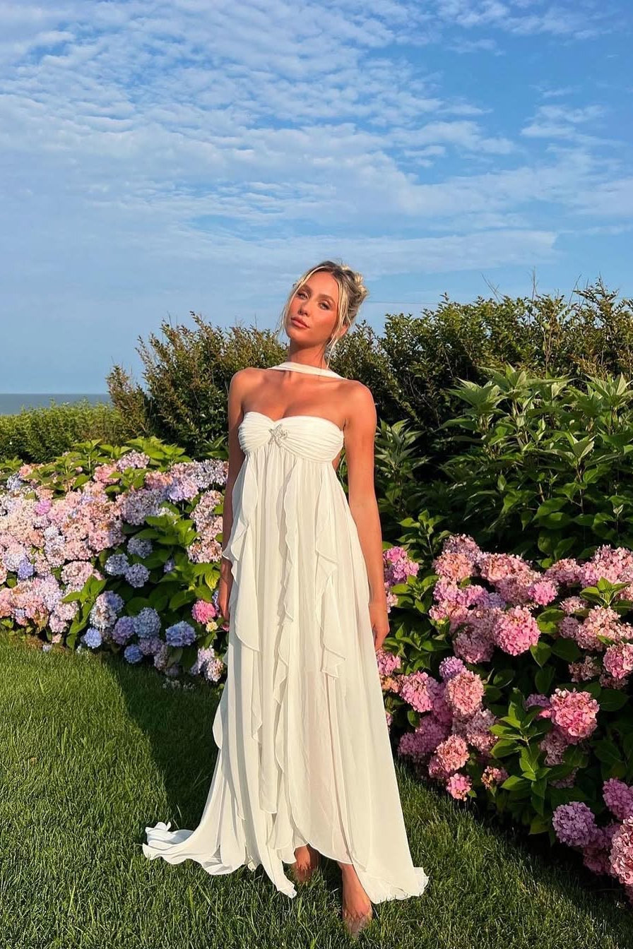 White strapless ruched ruffle maxi dress with a starfish brooch and choker, standing in a vibrant flower garden.