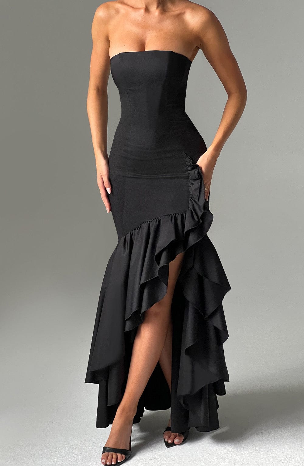 Close-up front view of a black strapless ruffle high-low maxi dress, emphasizing the leg slit and skirt ruffles.