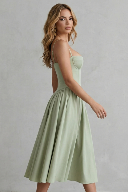 Side profile of a woman wearing a light sage green strappy structured sweetheart neck midi dress, showing the full pleated skirt.