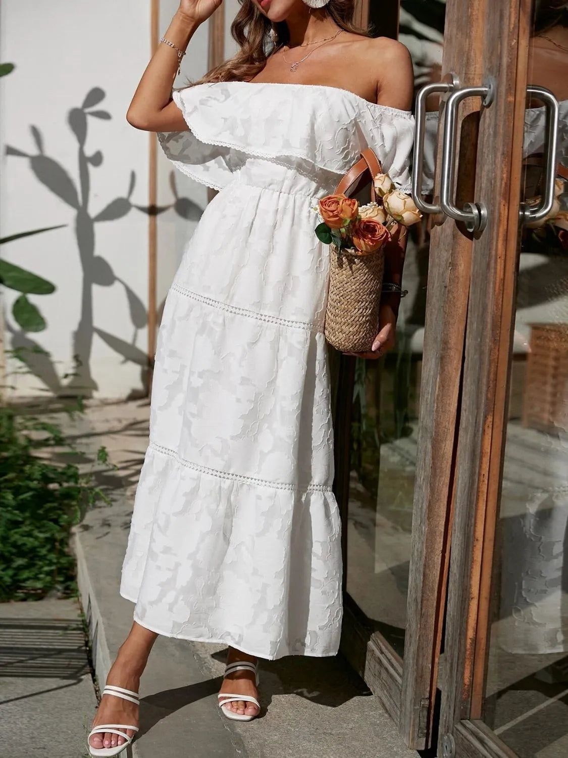 Elegant white off-shoulder tiered maxi dress with subtle floral texture, worn with a straw hat and white sandals, ideal for summer events.