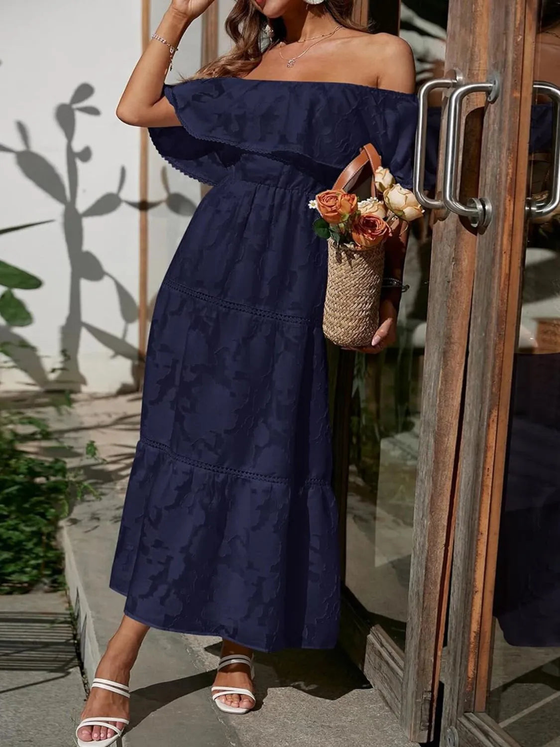 Navy blue off-shoulder tiered maxi dress for women, featuring a ruffled bodice and textured fabric, styled with white sandals and a straw bag.