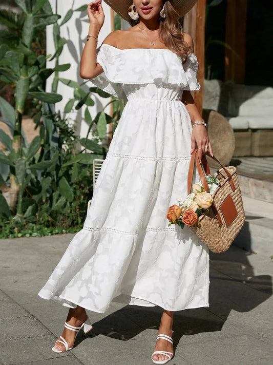 White off-shoulder tiered maxi dress for women, with ruffled neckline and textured fabric, paired with white block heels and a straw bag.